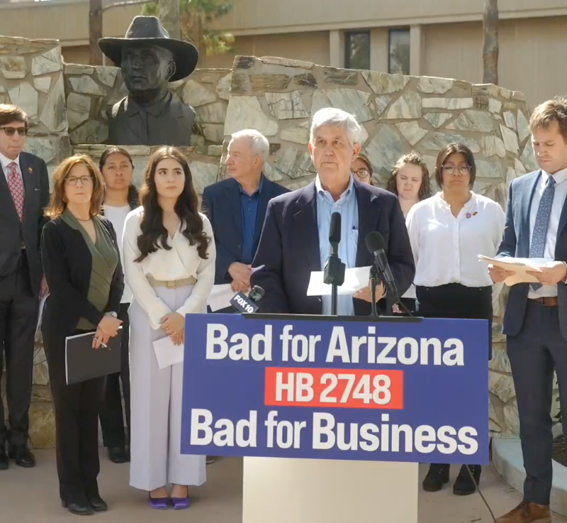 This image shows a group of diverse individuals standing behind a podium with a sign that reads 'Bad for Arizona HB 2748 Bad for Business' prominently displayed.